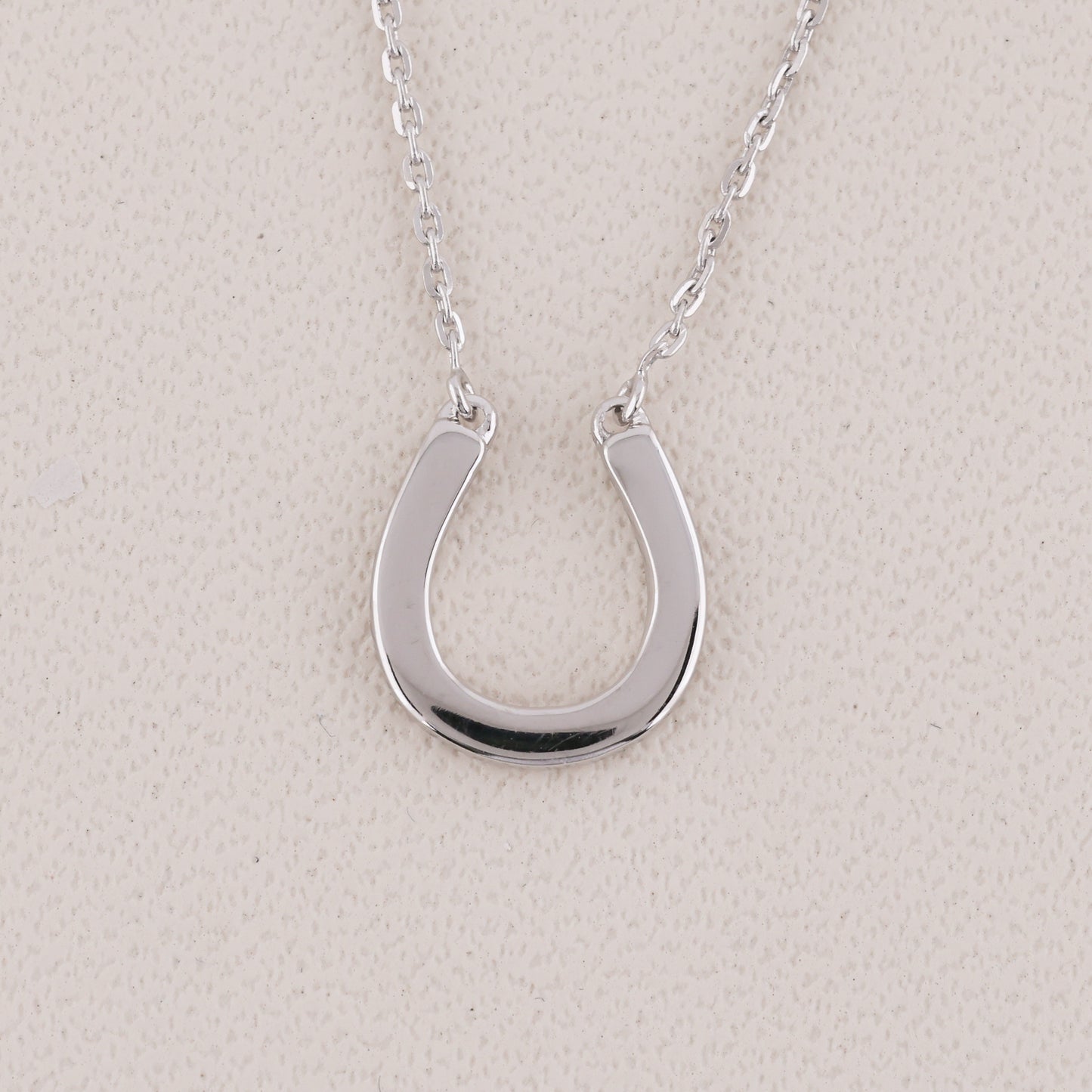 A polished silver horseshoe pendant necklace on a delicate silver chain, displayed against a textured light beige background.