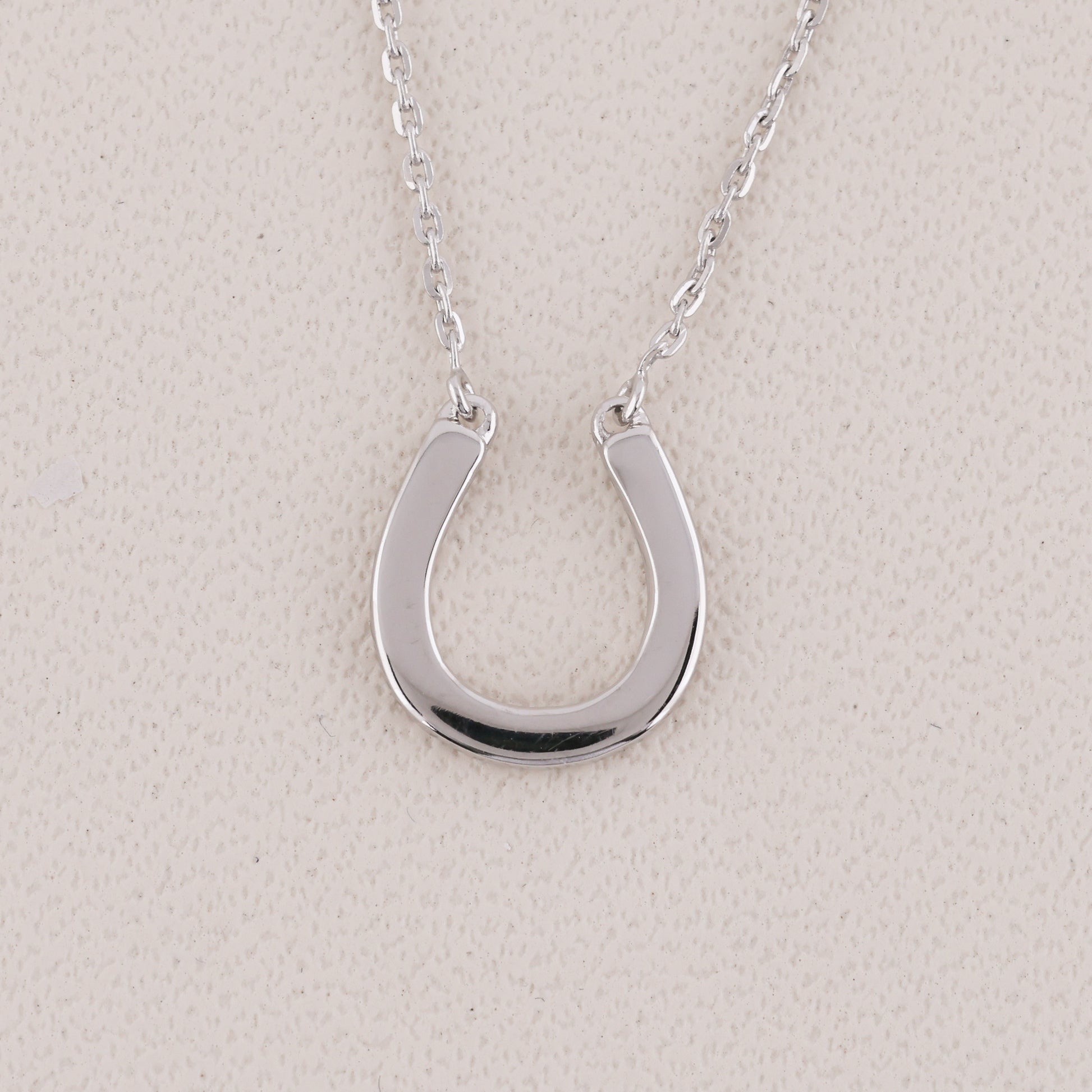 A polished silver horseshoe pendant necklace on a delicate silver chain, displayed against a textured light beige background.