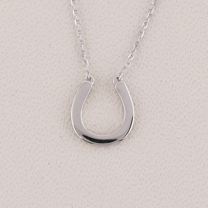 A polished silver horseshoe pendant necklace on a delicate silver chain, displayed against a textured light beige background.