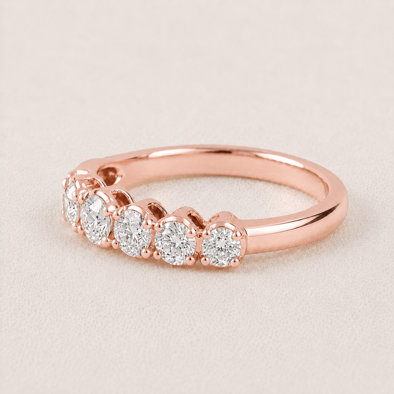 A rose gold diamond ring with five round-cut diamonds set in a row on a polished band, shown on a light beige textured background.