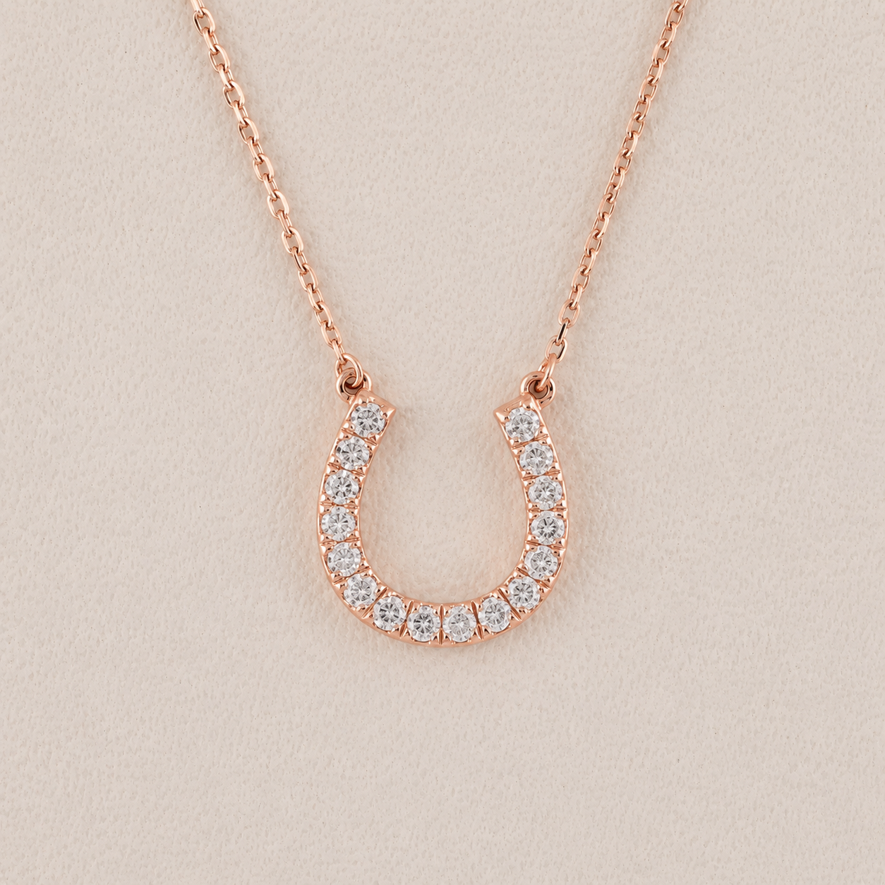 A rose gold horseshoe pendant necklace adorned with small clear stones, displayed on a textured beige background.
