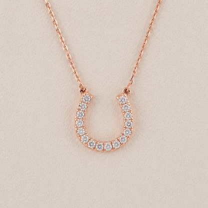 A rose gold horseshoe pendant necklace adorned with small clear stones, displayed on a textured beige background.