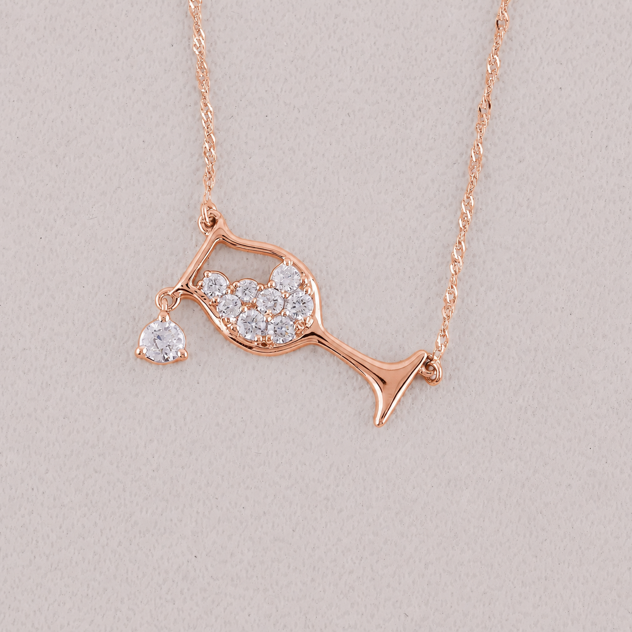 A rose gold necklace featuring a pendant shaped like a wine glass filled with small clear stones, with a dangling clear heart-shaped stone, displayed on a textured light beige background.