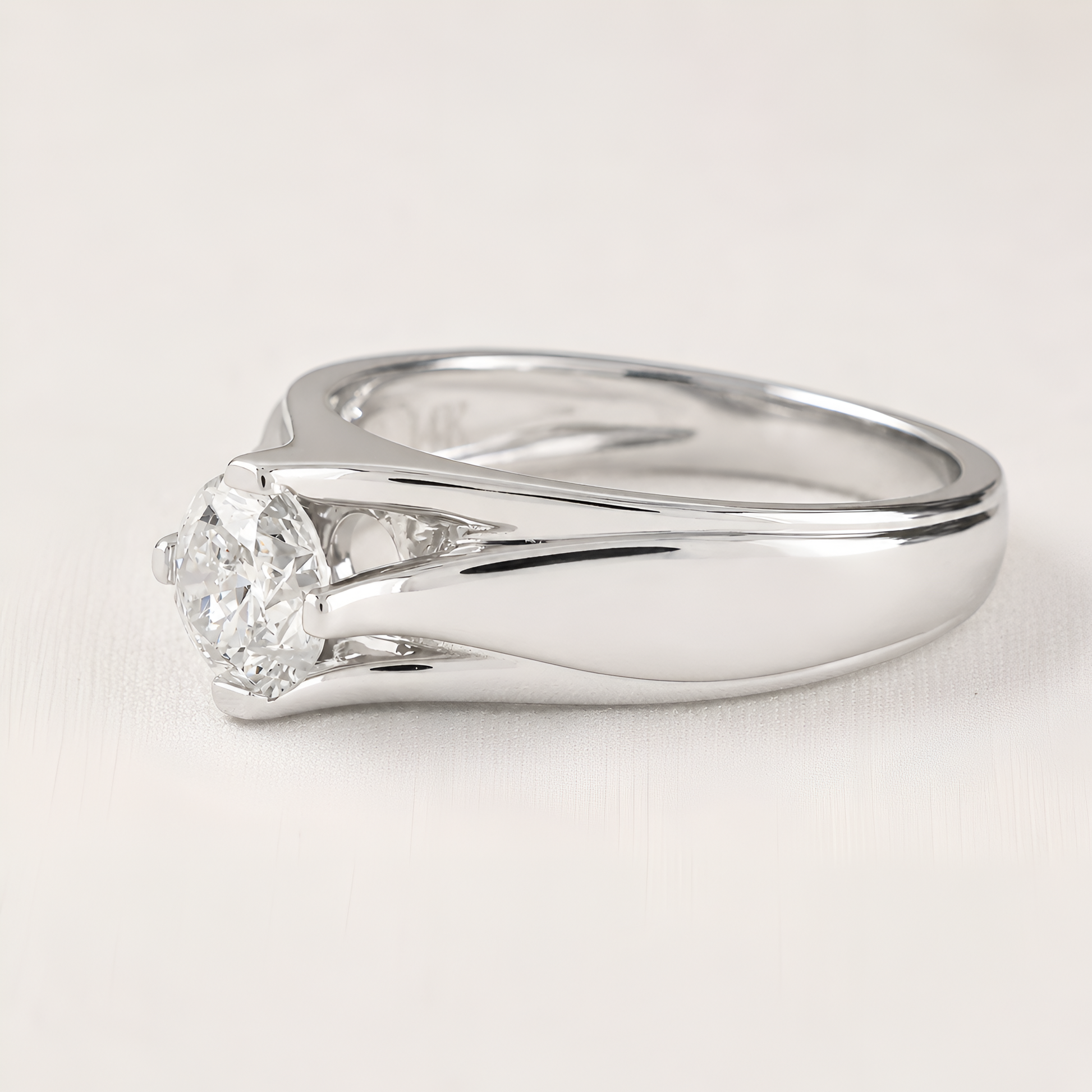 A silver-colored diamond ring with a round-cut center stone and a split shank design, shown on a light neutral background.
