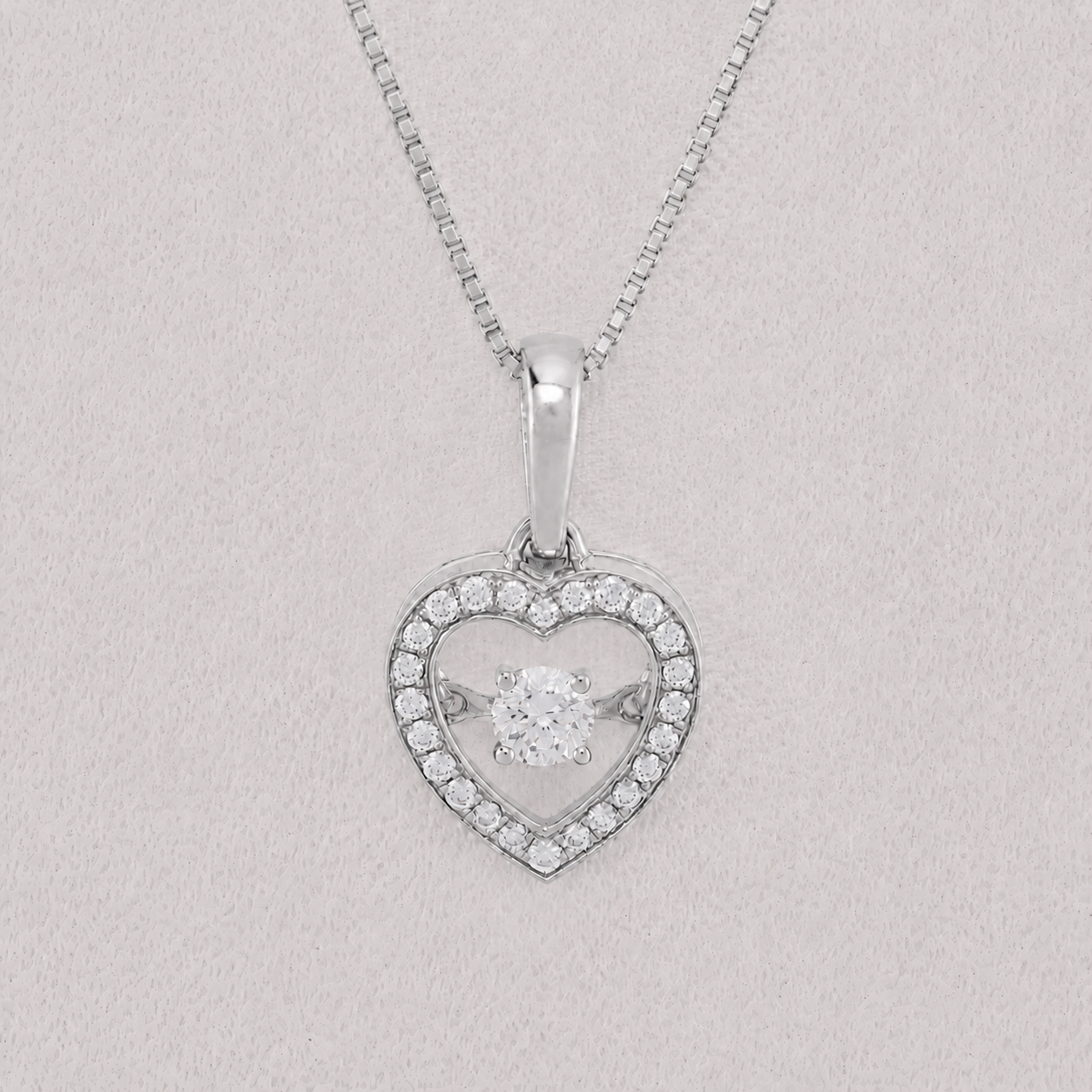 A silver heart-shaped pendant necklace with a halo border of small clear stones and a central round-cut stone, suspended from a delicate silver chain against a light gray textured background.