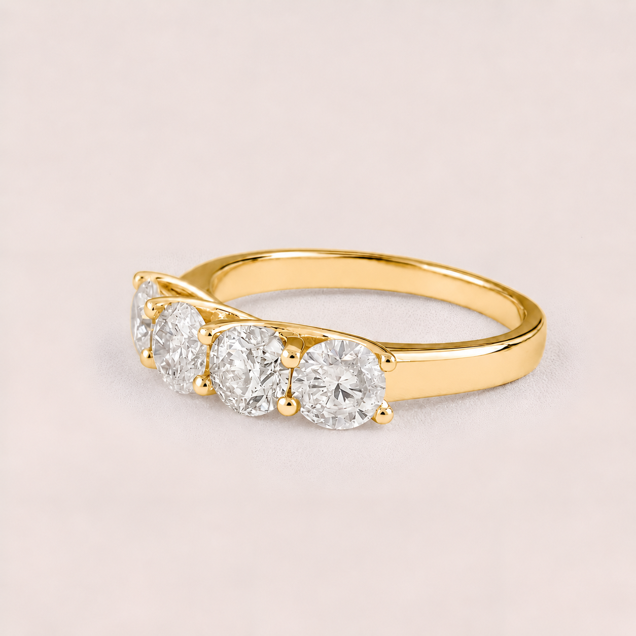 A yellow gold ring featuring three round-cut clear gemstones in a row, set on a plain light pink background.
