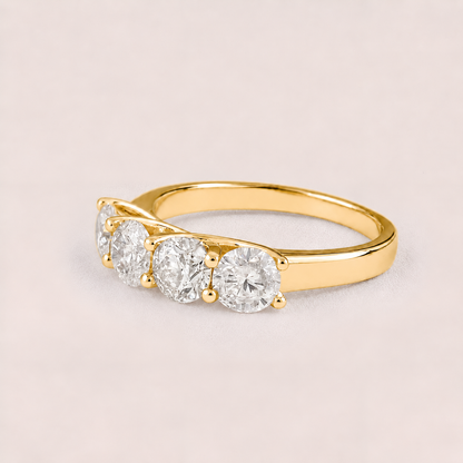 A yellow gold ring featuring three round-cut clear gemstones in a row, set on a plain light pink background.