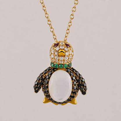 Penguin-shaped pendant with gemstones on a gold chain against a beige background