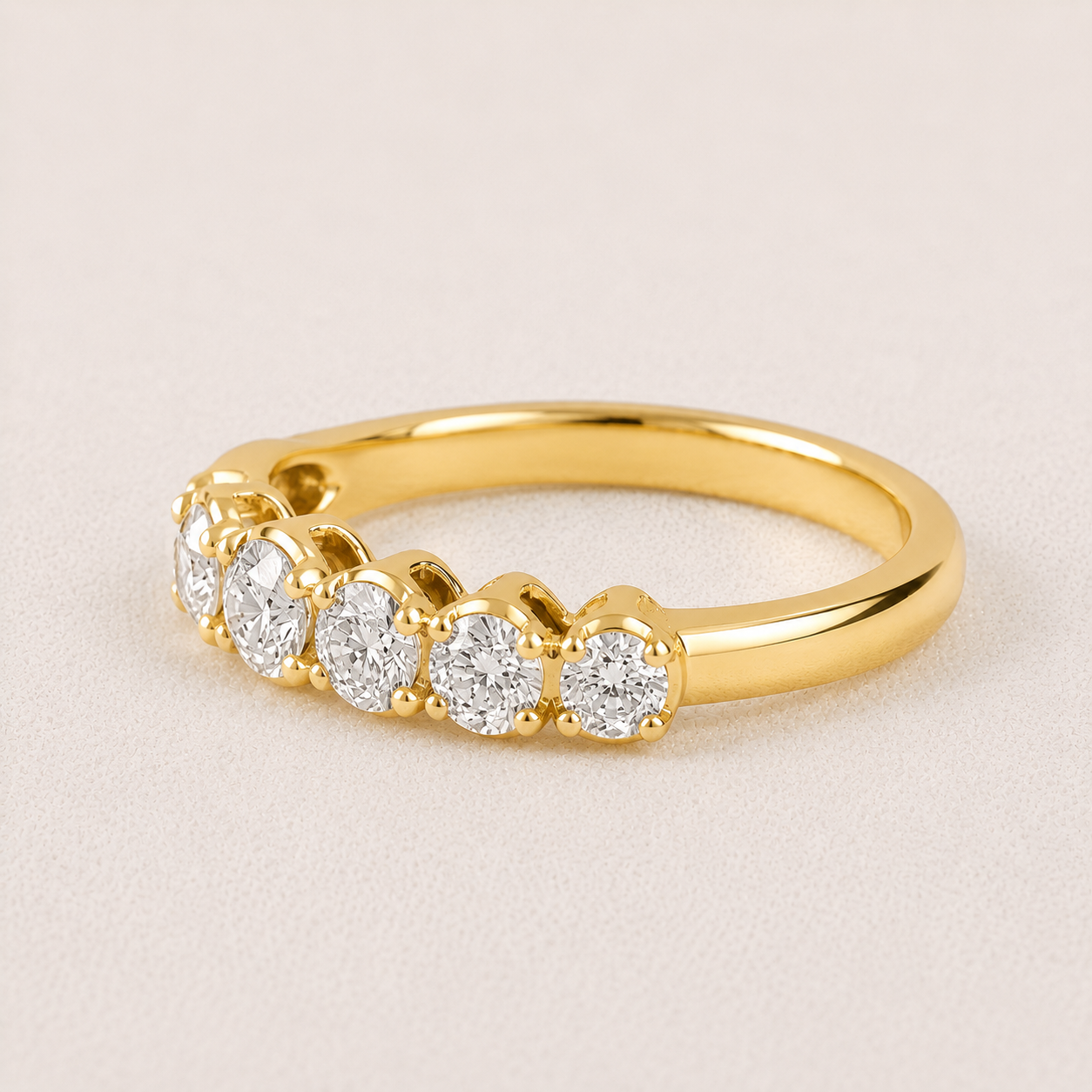 A yellow gold diamond ring with five round-cut diamonds set in a row, shown on a light beige textured background.