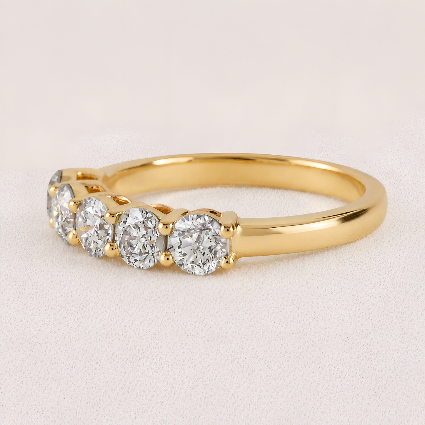 A yellow gold five-stone diamond ring with round-cut diamonds set in a prong setting, shown on a light beige textured background.