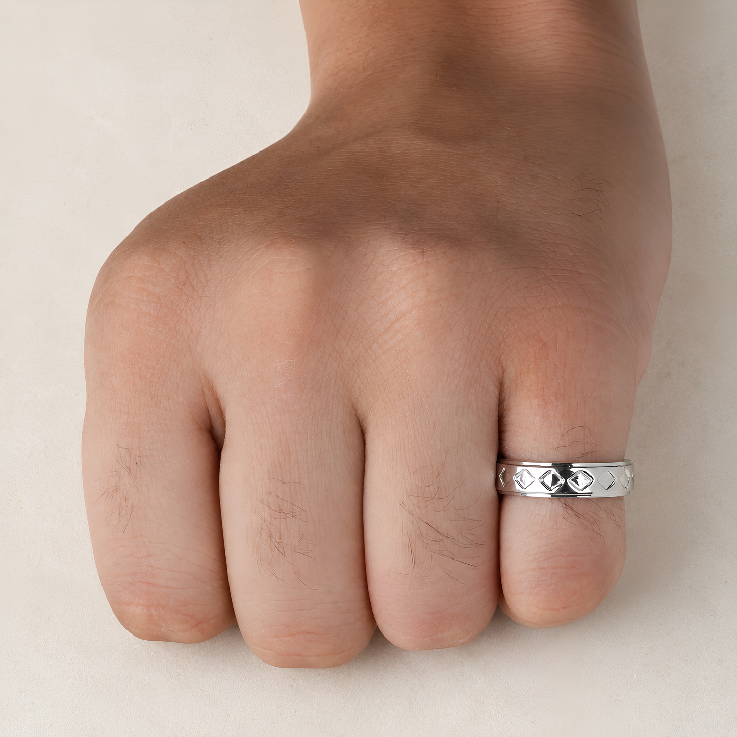 A silver band ring with a textured, diamond-like pattern worn on the ring finger of a hand, shown against a plain light beige background.