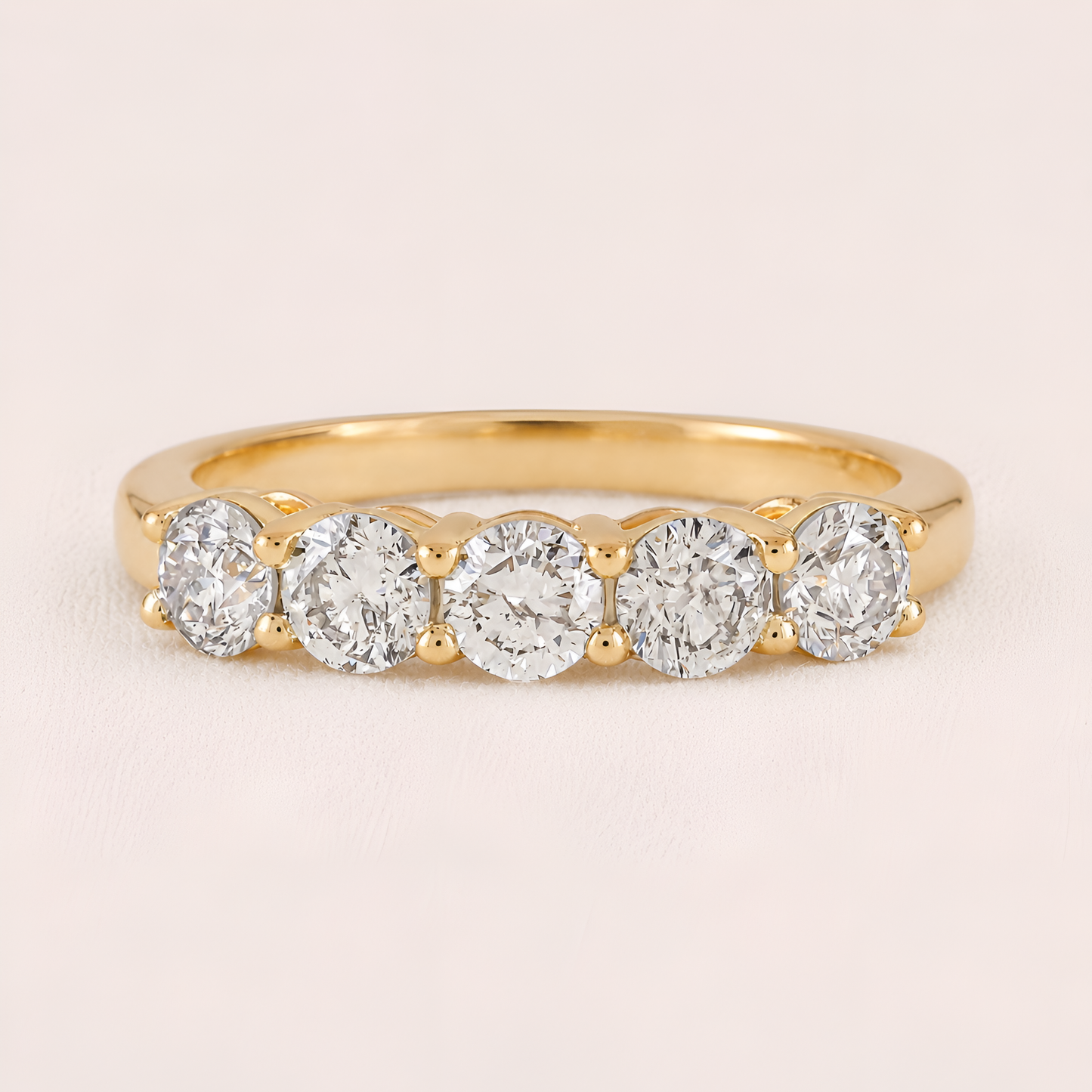 A yellow gold five-stone diamond ring with round-cut diamonds set in a row on a polished band, shown against a light pink background.