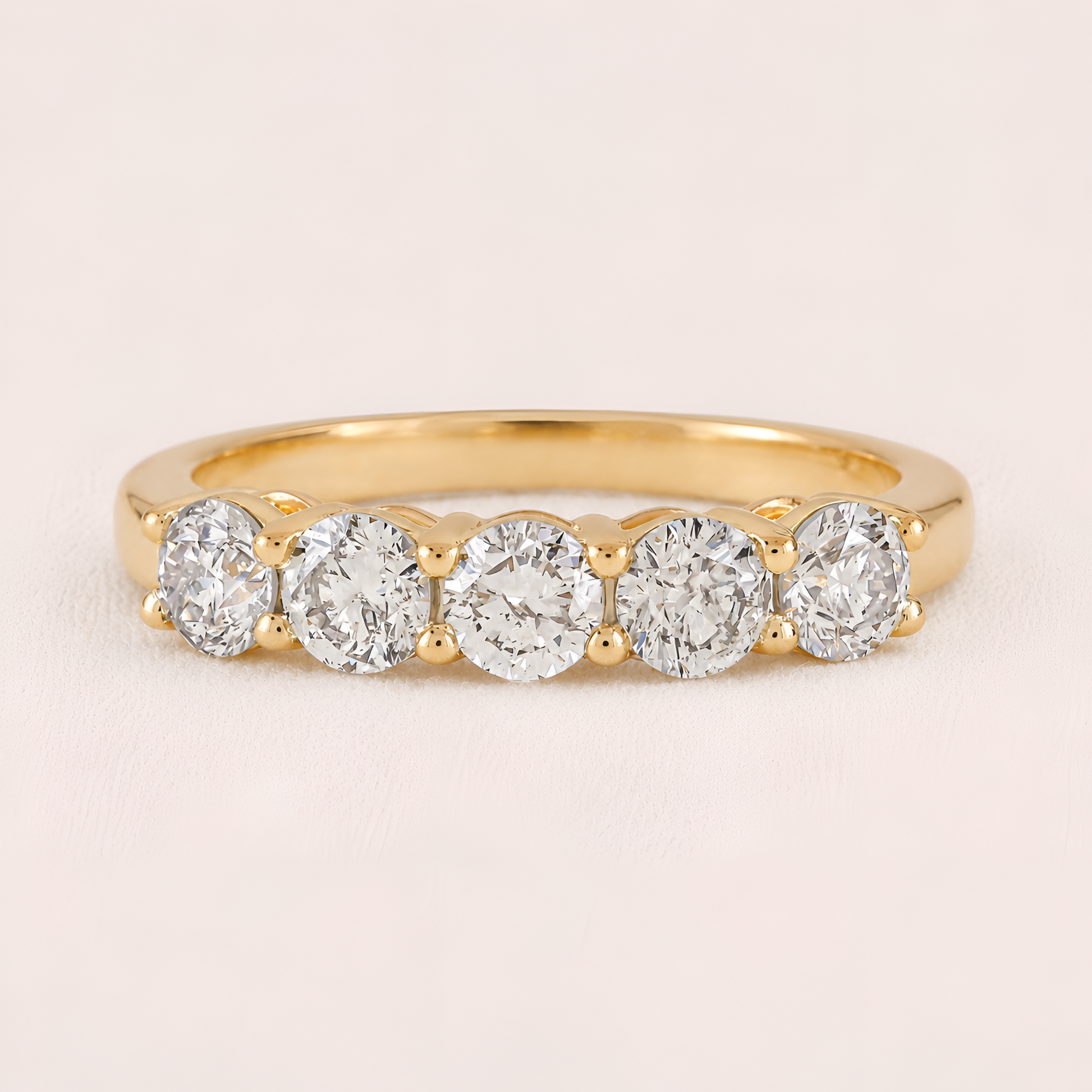 A yellow gold five-stone diamond ring with round-cut diamonds set in a row on a polished band, shown against a light pink background.
