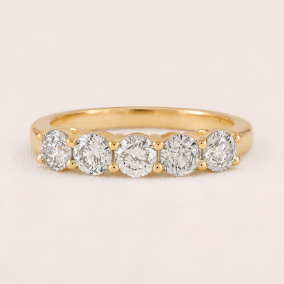 A yellow gold five-stone diamond ring with round-cut diamonds set in a row on a polished band, shown against a light pink background.