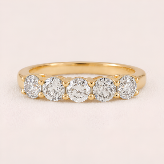 A yellow gold five-stone diamond ring with round-cut diamonds set in a row on a polished band, shown against a light pink background.