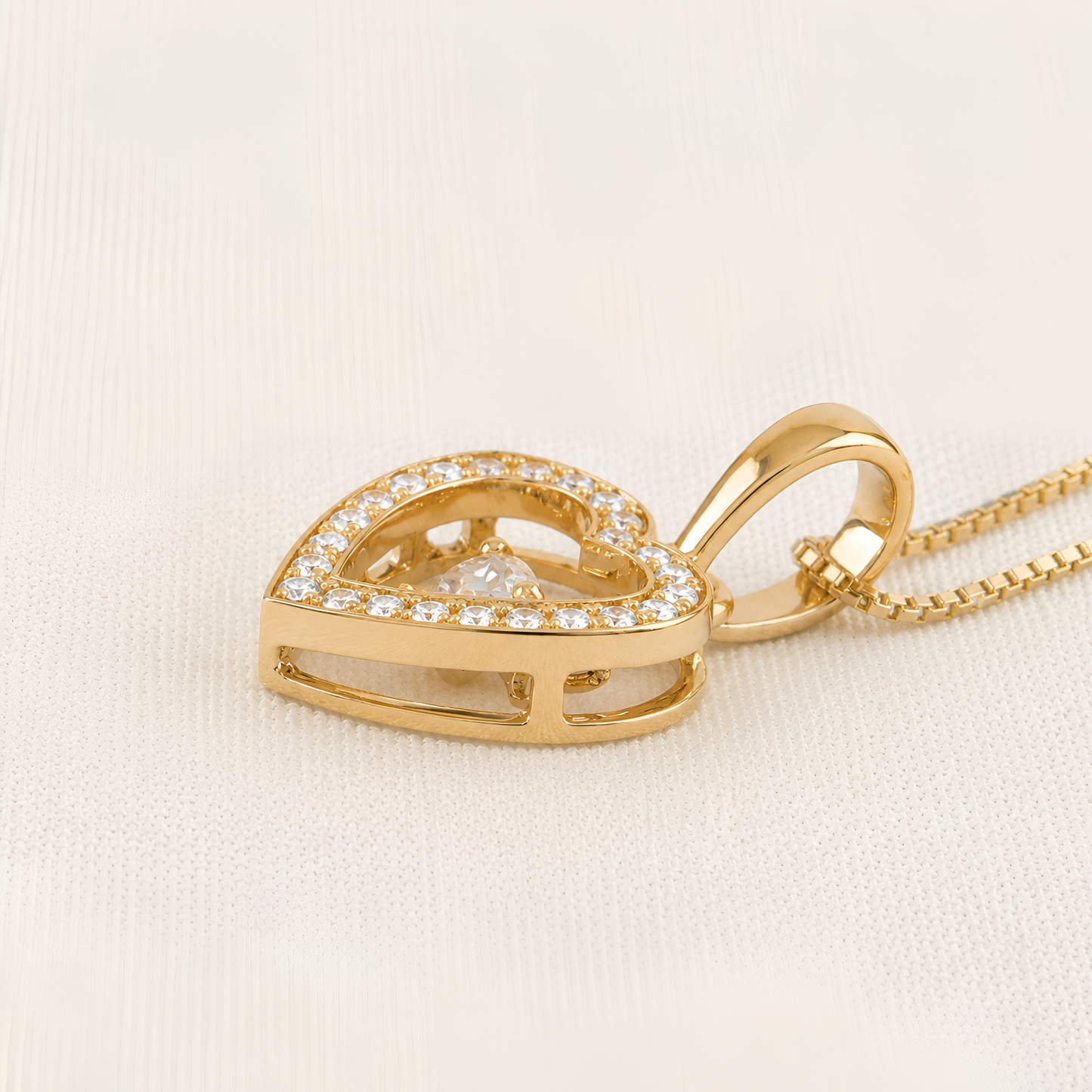 A gold heart-shaped pendant necklace with a double row of small clear stones and a central clear gemstone, shown on a textured off-white background.