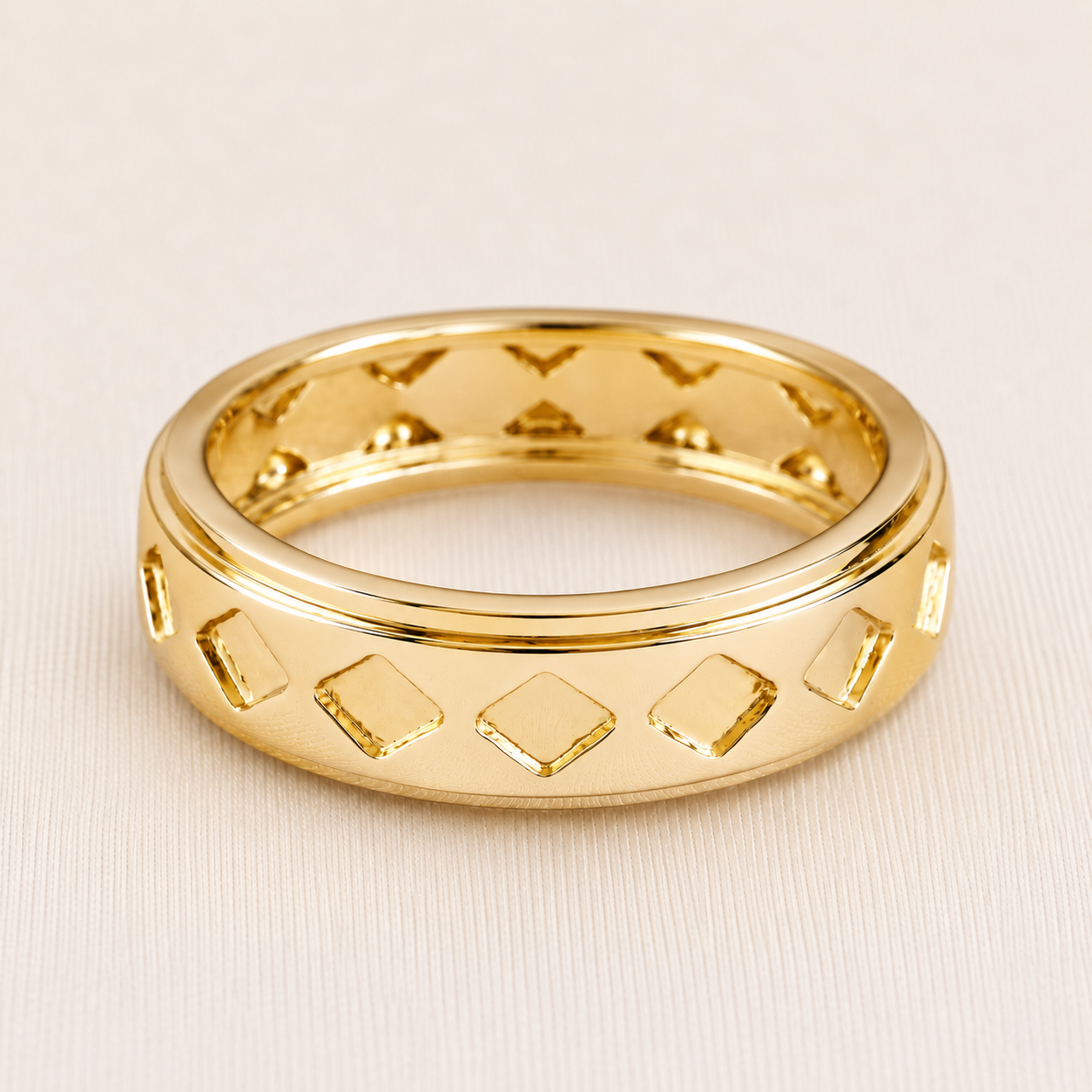 A polished gold band ring with a geometric pattern of raised and recessed diamond shapes, photographed on a light beige textured background.