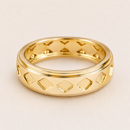 A polished gold band ring with a geometric pattern of raised and recessed diamond shapes, photographed on a light beige textured background.