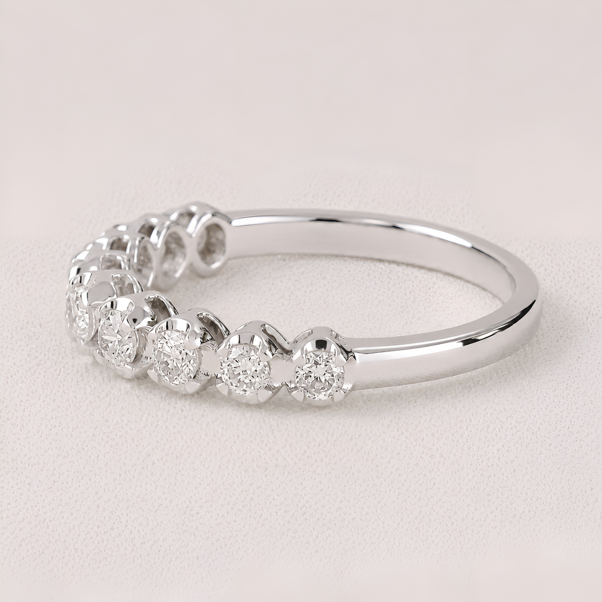 A silver diamond eternity ring featuring a row of round-cut diamonds in a halo setting on a polished band, photographed on a light beige textured background.
