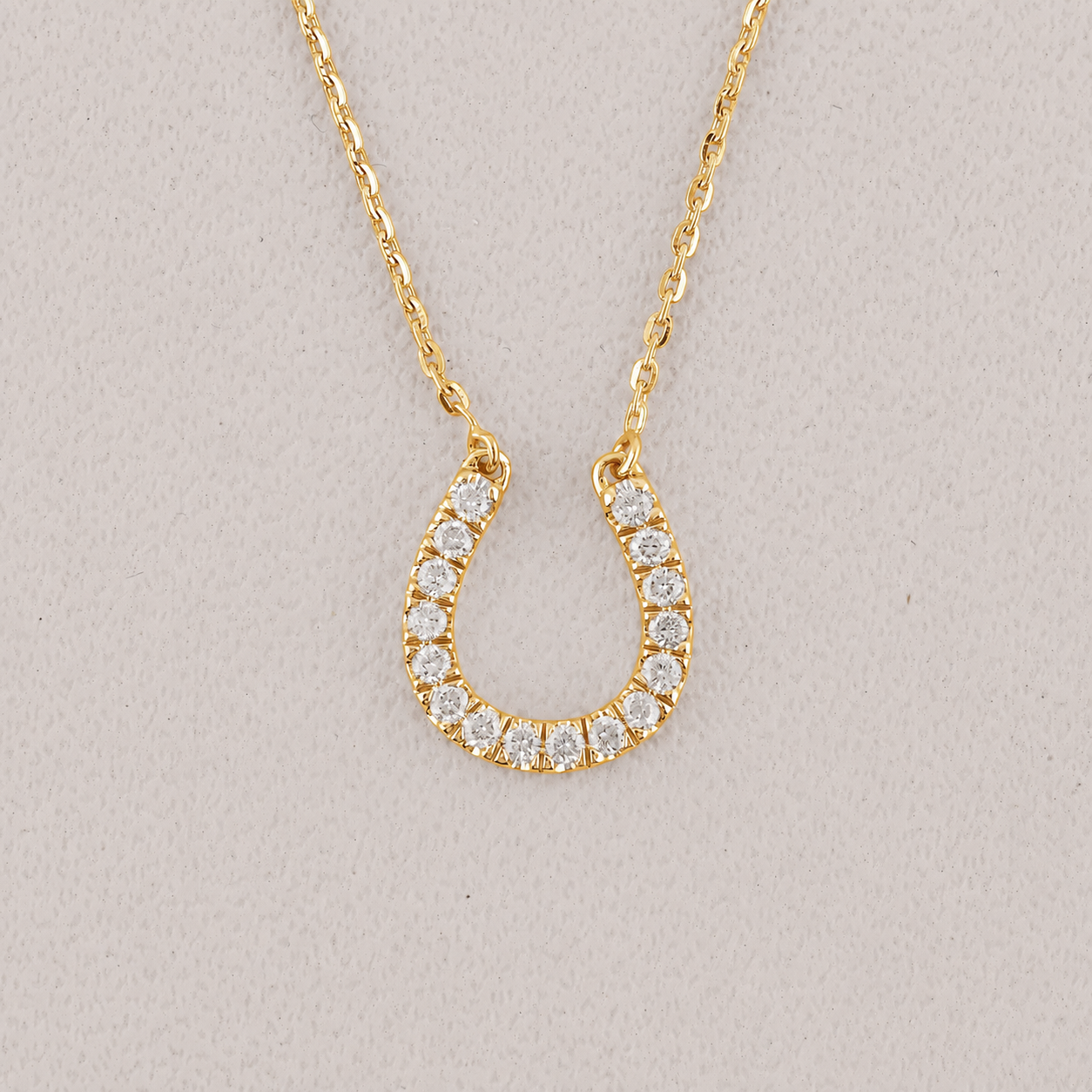 A gold horseshoe-shaped pendant necklace adorned with small clear stones, displayed on a textured light gray background.
