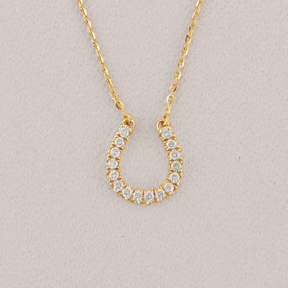A gold horseshoe-shaped pendant necklace adorned with small clear stones, displayed on a textured light gray background.