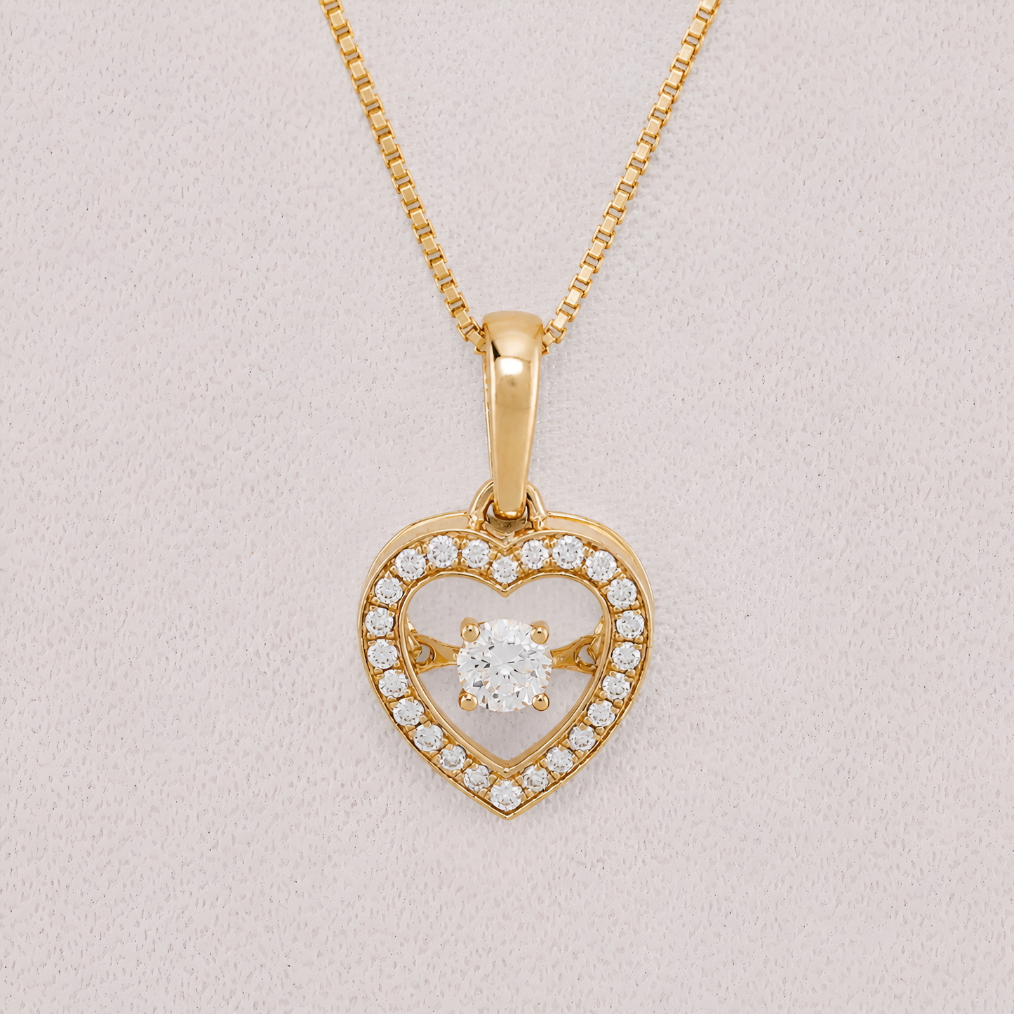A yellow gold heart-shaped pendant necklace with a central round-cut clear gemstone and a border of small pavé-set clear stones, shown on a textured light pink background.