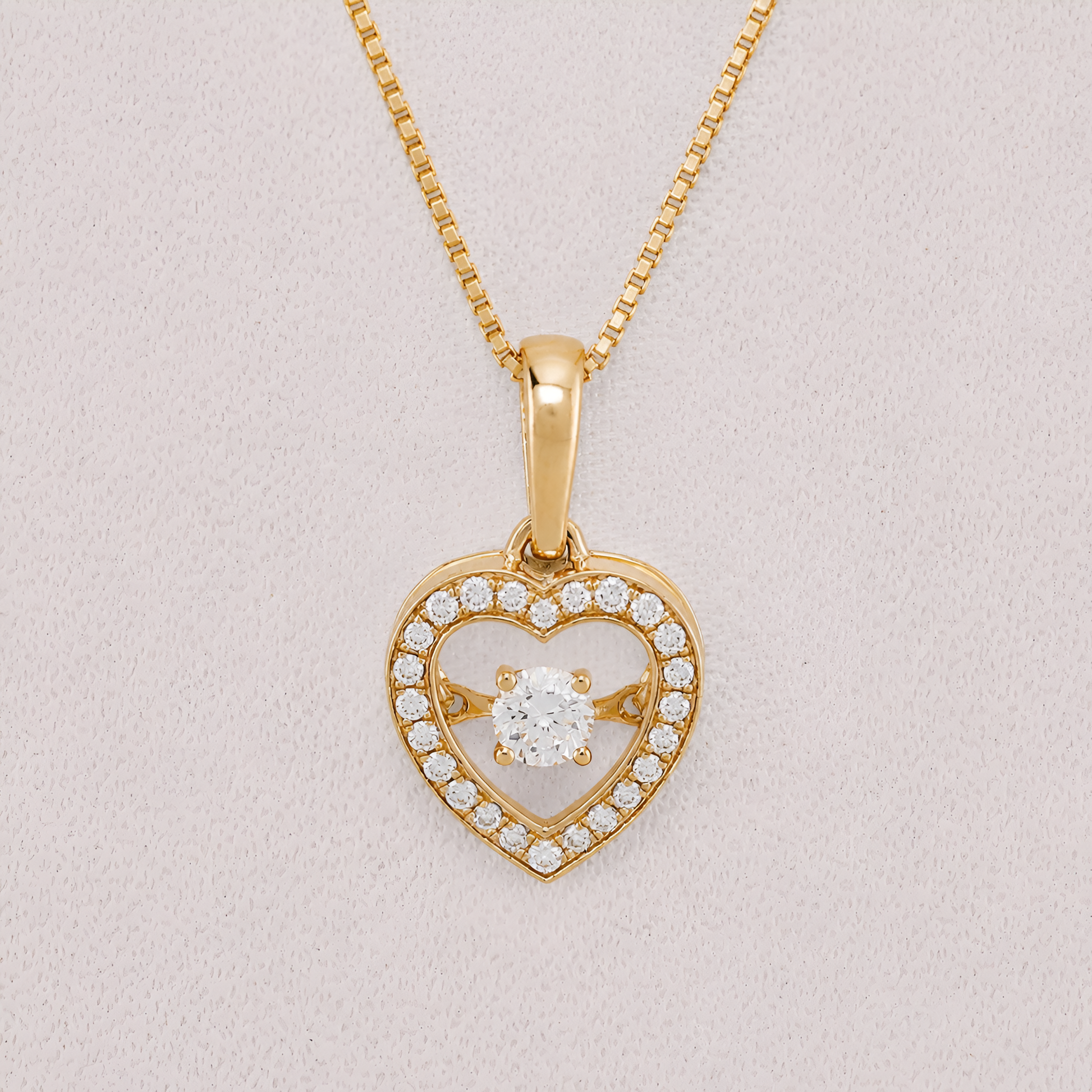 A yellow gold heart-shaped pendant necklace with a central round-cut clear gemstone and a border of small pavé-set clear stones, shown on a textured light pink background.