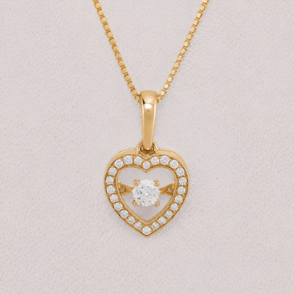 A yellow gold heart-shaped pendant necklace with a central round-cut clear gemstone and a border of small pavé-set clear stones, shown on a textured light pink background.