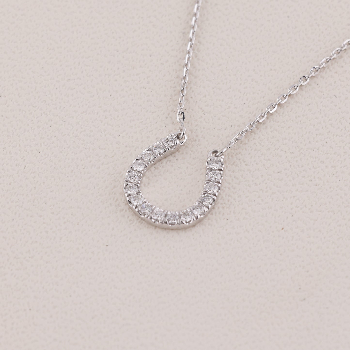 A silver horseshoe-shaped pendant necklace encrusted with small clear stones, shown on a delicate silver chain against a light beige textured background.
