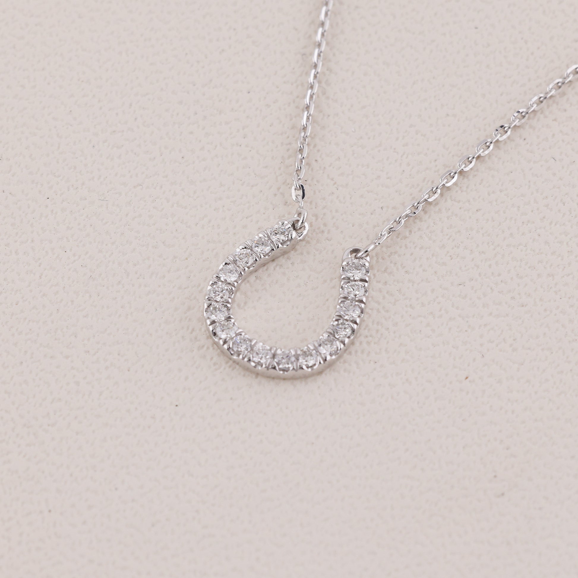 A silver horseshoe-shaped pendant necklace encrusted with small clear stones, shown on a delicate silver chain against a light beige textured background.