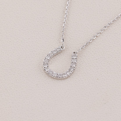 A silver horseshoe-shaped pendant necklace encrusted with small clear stones, shown on a delicate silver chain against a light beige textured background.