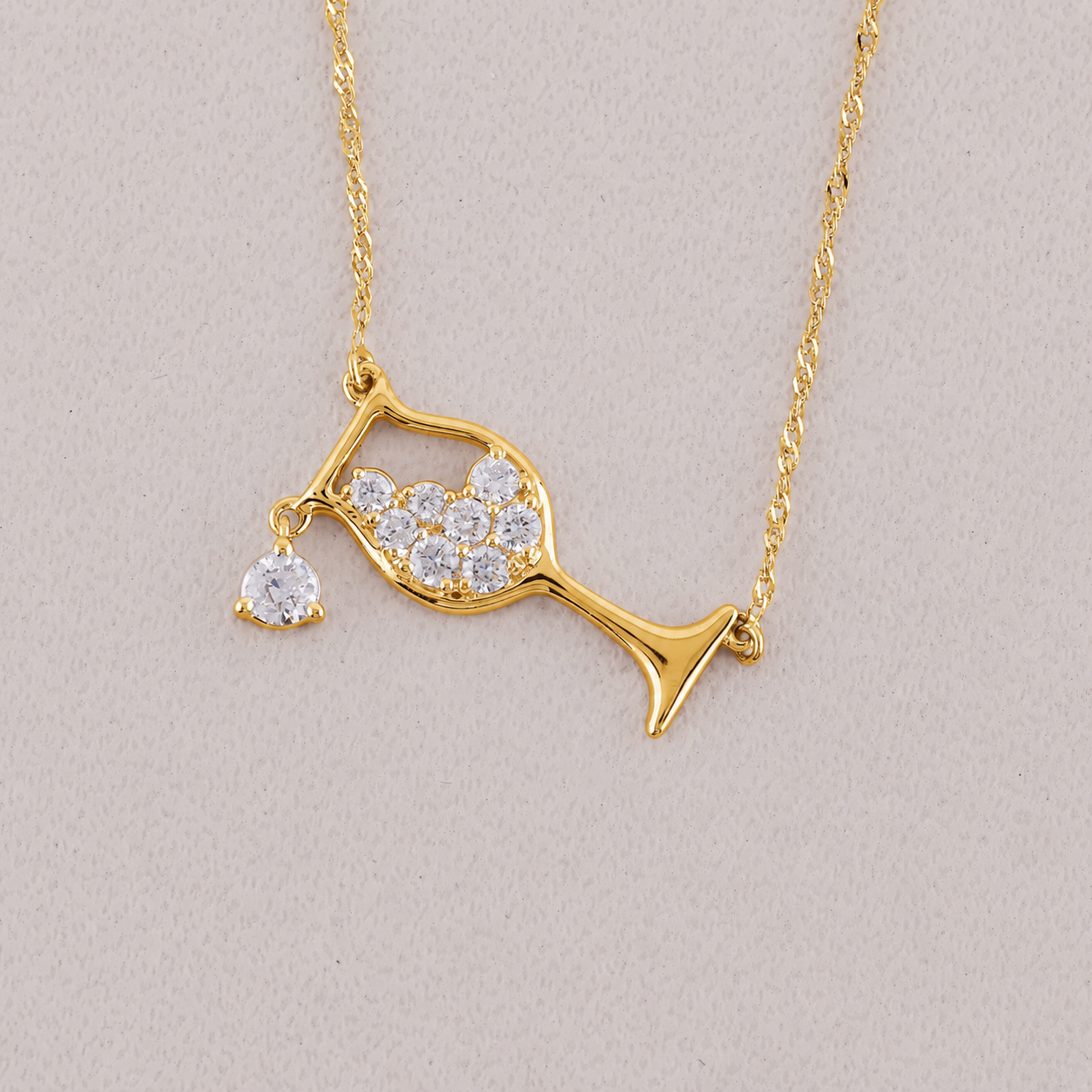 A gold necklace featuring a wine glass pendant adorned with clear pavé stones and a dangling heart-shaped clear stone, displayed on a neutral light beige background.