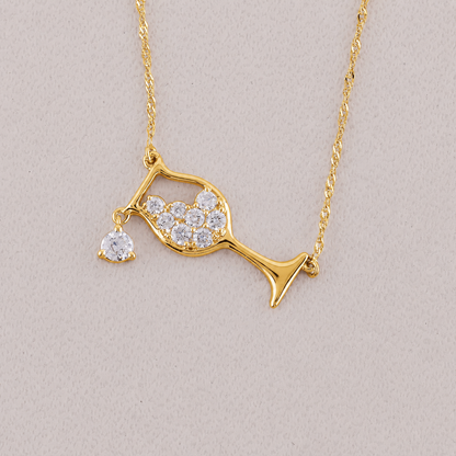 A gold necklace featuring a wine glass pendant adorned with clear pavé stones and a dangling heart-shaped clear stone, displayed on a neutral light beige background.