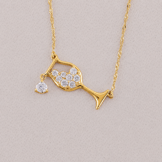A gold necklace featuring a wine glass pendant adorned with clear pavé stones and a dangling heart-shaped clear stone, displayed on a neutral light beige background.