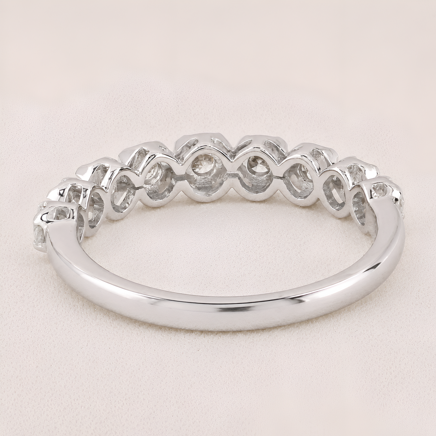 A silver band ring featuring a row of small, bezel-set clear gemstones in a repeating wavy pattern, shown on a light beige textured background.
