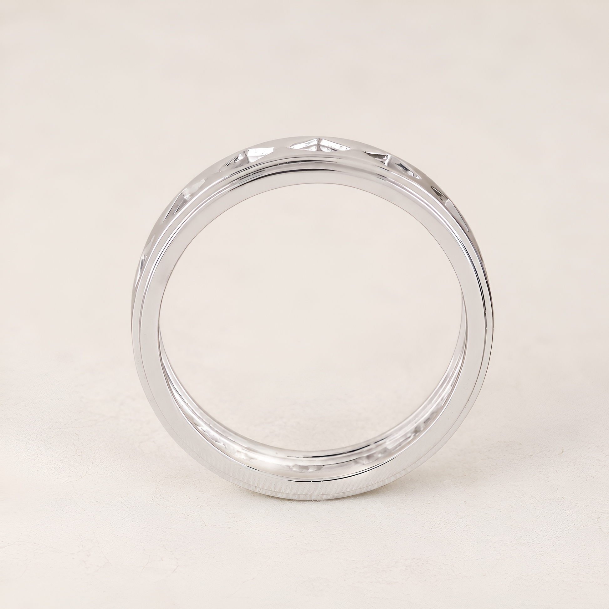 A polished silver band ring with a beveled edge and a subtle textured pattern on the outer surface, displayed on a light beige background.
