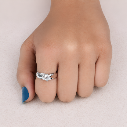 A silver ring with a clear heart-shaped gemstone worn on a person's ring finger, shown against a soft beige background.