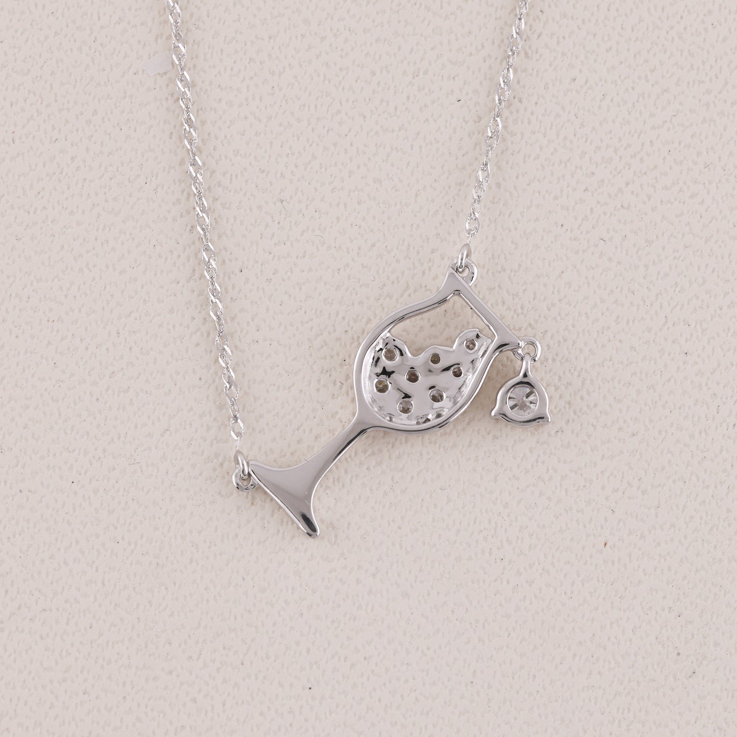 A silver necklace featuring a wine glass pendant with a heart-shaped cutout and a small dangling heart charm, displayed on a textured light beige background.