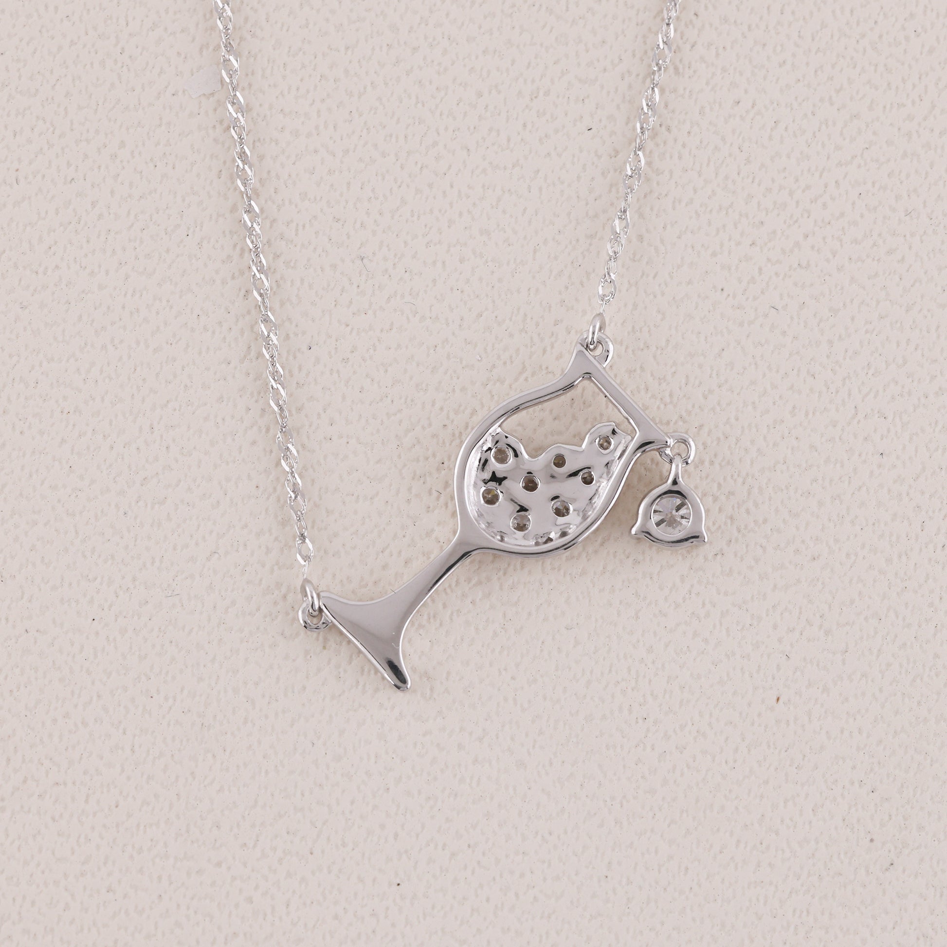A silver necklace featuring a wine glass pendant with a heart-shaped cutout and a small dangling heart charm, displayed on a textured light beige background.