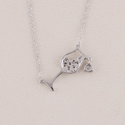 A silver necklace featuring a wine glass pendant with a heart-shaped cutout and a small dangling heart charm, displayed on a textured light beige background.
