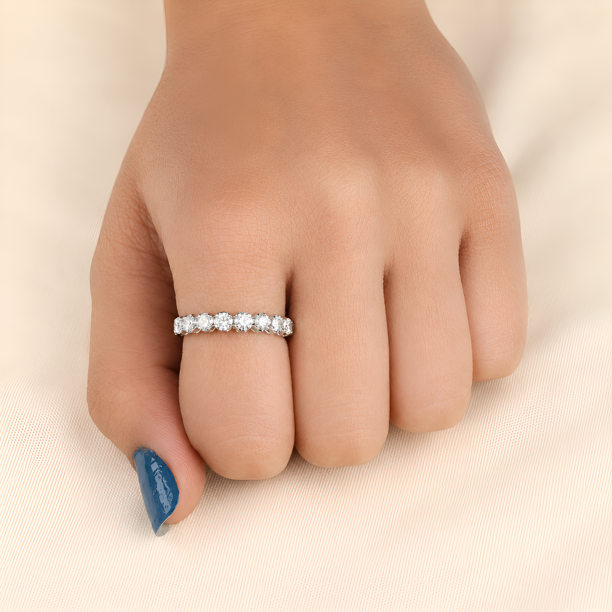 A close-up of a hand wearing a thin silver eternity ring set with small clear stones, resting on a soft beige fabric background.