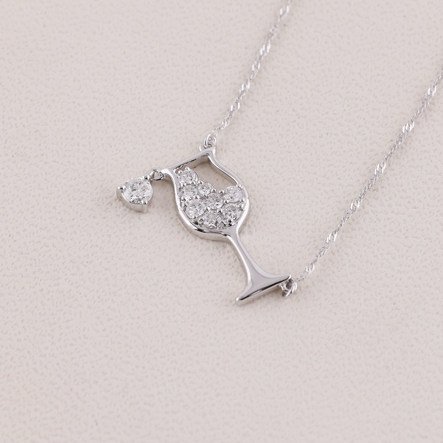 A silver necklace featuring a wine glass pendant paved with clear stones and a small heart-shaped pendant, displayed on a textured light beige surface.