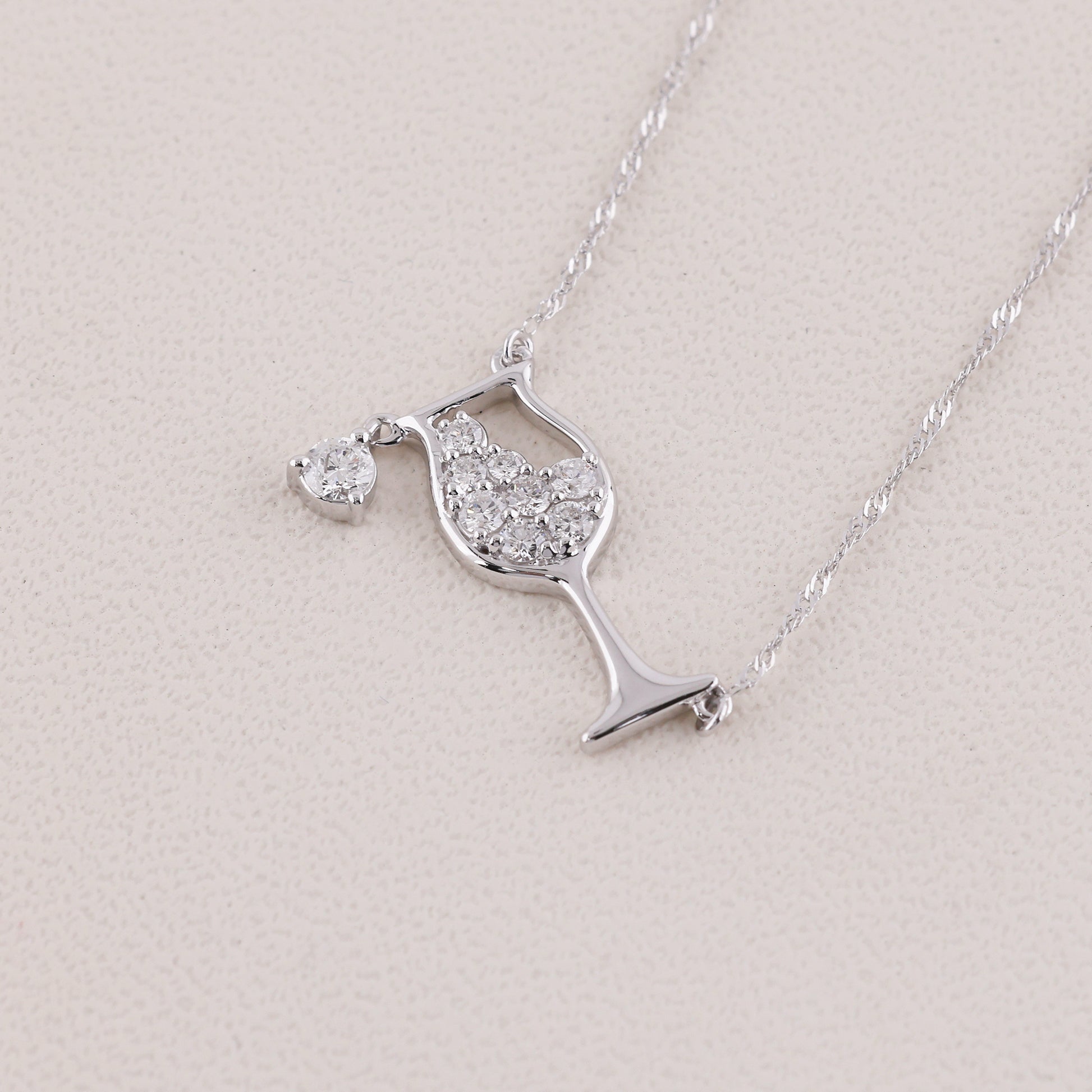 A silver necklace featuring a wine glass pendant paved with clear stones and a small heart-shaped pendant, displayed on a textured light beige surface.