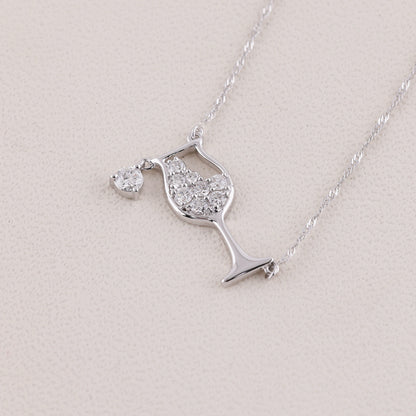 A silver necklace featuring a wine glass pendant paved with clear stones and a small heart-shaped pendant, displayed on a textured light beige surface.
